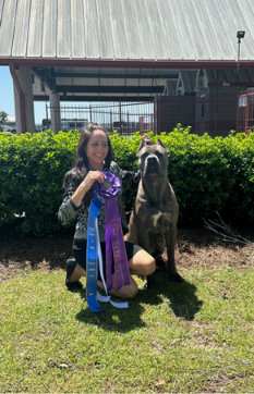 Woman with dog holding award ribbons outside.